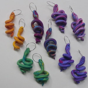 May include: Eight pairs of handmade polymer clay earrings shaped like snakes. The snakes are in various colours, including orange, green, purple, and blue. The earrings are made with silver hoops.