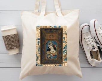 Puede incluir: Un bolso de tela color crema con un gráfico rectangular central de una persona con cabello oscuro en un marco decorativo dorado y azul, rodeado por un patrón floral. El bolso tiene dos correas para el hombro y una estructura de tela sencilla.