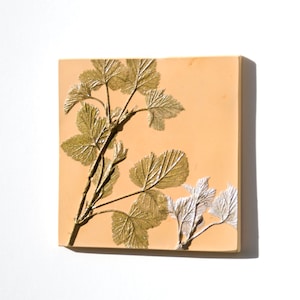 Puede incluir: Azulejo cuadrado de color melocotón con un diseño botánico en relieve. El diseño presenta una rama con hojas detalladas en tonos dorados y plateados. El azulejo está montado en una pared blanca.
