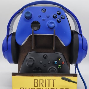 May include: A wooden stand with a blue and black Xbox controller and a blue headset. The stand has a yellow sign that says "Britt Chronicles".