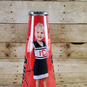 Peut inclure: Un mégaphone de cheerleading rouge et blanc avec une photo d'une jeune fille portant un uniforme de cheerleading noir et blanc avec les lettres "FCS" sur le devant. Le mégaphone a un sommet argenté et un fond rouge craquelé.