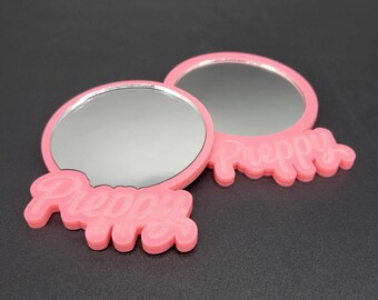 Peut inclure: Deux miroirs compacts roses avec le mot "Preppy" en relief sur le bas. Les miroirs sont ronds et ont une surface réfléchissante argentée.