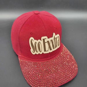 May include: A red baseball cap with a brown leather patch that says "Soo Exxtra" on the front. The brim of the cap is covered in rhinestones.