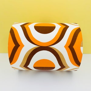 Puede incluir: Un coj&iacute;n cil&iacute;ndrico con un patr&oacute;n geom&eacute;trico retro. El dise&ntilde;o presenta formas curvas repetidas en tonos naranja, marr&oacute;n y beige sobre un fondo blanco. El coj&iacute;n es un art&iacute;culo decorativo.