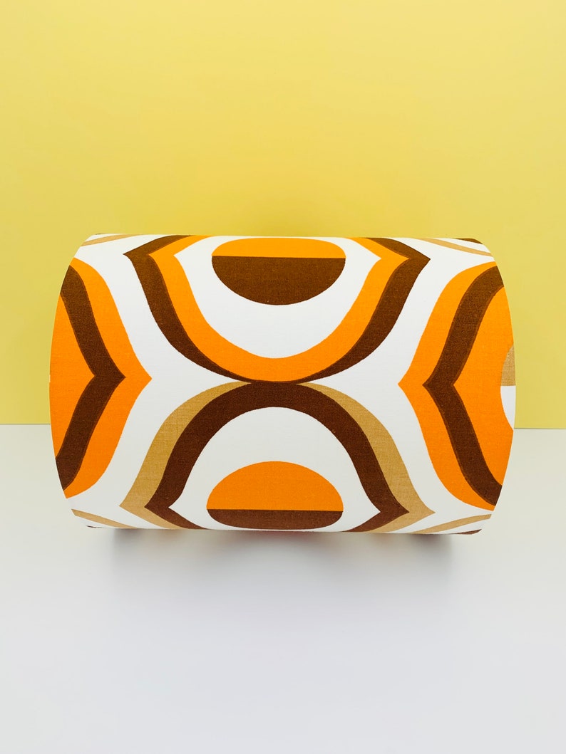Puede incluir: Un coj&iacute;n cil&iacute;ndrico con un patr&oacute;n geom&eacute;trico retro. El dise&ntilde;o presenta formas curvas repetidas en tonos naranja, marr&oacute;n y beige sobre un fondo blanco. El coj&iacute;n es un art&iacute;culo decorativo.