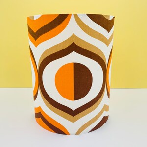Puede incluir: Pantalla de l&aacute;mpara cil&iacute;ndrica con dise&ntilde;o retro. La tela presenta un patr&oacute;n repetitivo de l&iacute;neas y c&iacute;rculos curvos blancos, marrones, tostados y naranjas. La pantalla de la l&aacute;mpara est&aacute; sobre un fondo amarillo.