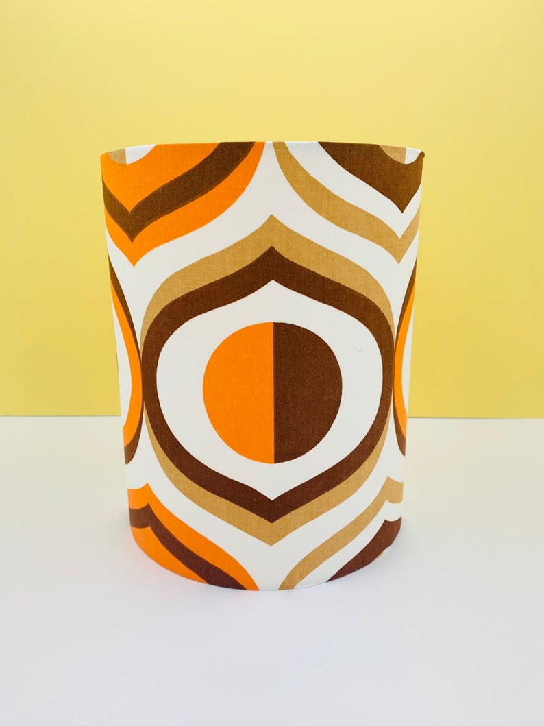 Puede incluir: Pantalla de l&aacute;mpara cil&iacute;ndrica con dise&ntilde;o retro. La tela presenta un patr&oacute;n repetitivo de l&iacute;neas y c&iacute;rculos curvos blancos, marrones, tostados y naranjas. La pantalla de la l&aacute;mpara est&aacute; sobre un fondo amarillo.