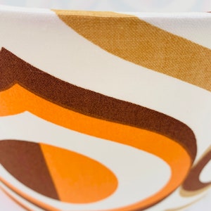 Puede incluir: Primer plano de una pantalla de l&aacute;mpara con dise&ntilde;o retro. La tela presenta un patr&oacute;n de rayas curvas en tonos marr&oacute;n, naranja y crema. La pantalla tiene forma redondeada y un marco de metal blanco.