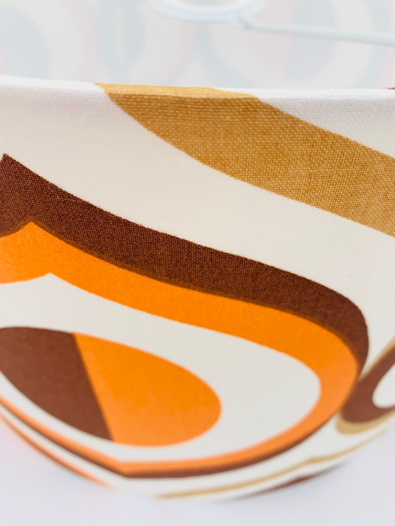 Puede incluir: Primer plano de una pantalla de l&aacute;mpara con dise&ntilde;o retro. La tela presenta un patr&oacute;n de rayas curvas en tonos marr&oacute;n, naranja y crema. La pantalla tiene forma redondeada y un marco de metal blanco.