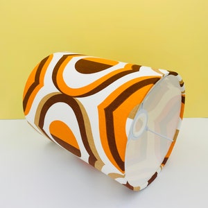 Puede incluir: Una pantalla de l&aacute;mpara cil&iacute;ndrica con un dise&ntilde;o retro. La pantalla presenta un patr&oacute;n repetitivo de l&iacute;neas curvas naranjas, marrones y blancas. El interior de la pantalla es blanco y el marco de metal es visible.