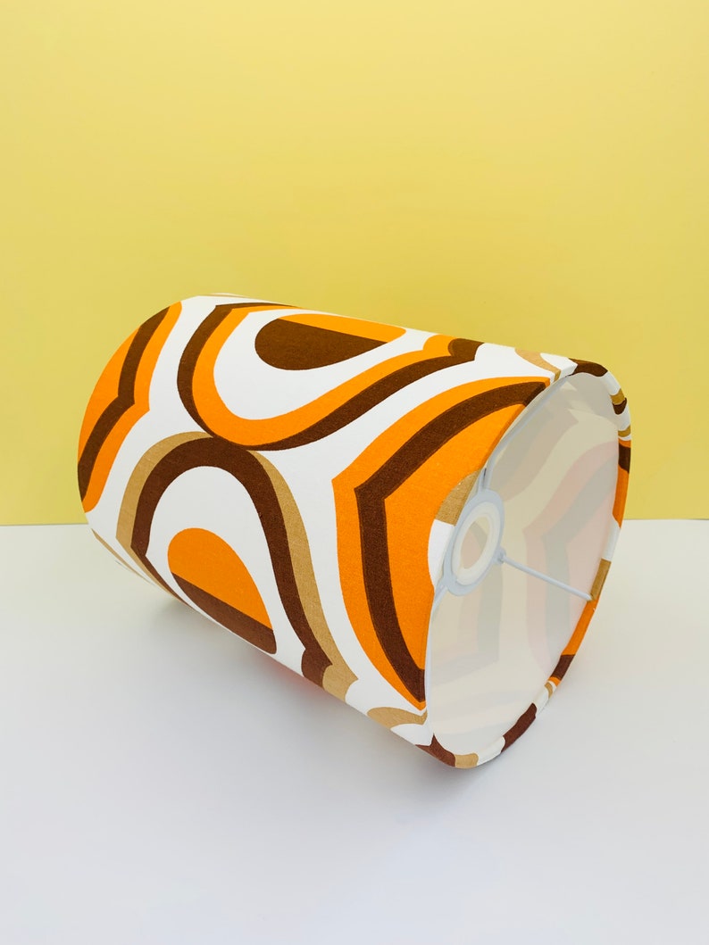 Puede incluir: Una pantalla de l&aacute;mpara cil&iacute;ndrica con un dise&ntilde;o retro. La pantalla presenta un patr&oacute;n repetitivo de l&iacute;neas curvas naranjas, marrones y blancas. El interior de la pantalla es blanco y el marco de metal es visible.