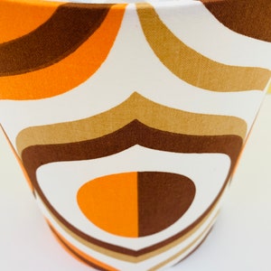 Puede incluir: Pantalla de l&aacute;mpara con dise&ntilde;o retro. La tela presenta un patr&oacute;n repetido de formas curvas en tonos marr&oacute;n, naranja y beige sobre un fondo blanco. La pantalla tiene una estructura met&aacute;lica blanca.