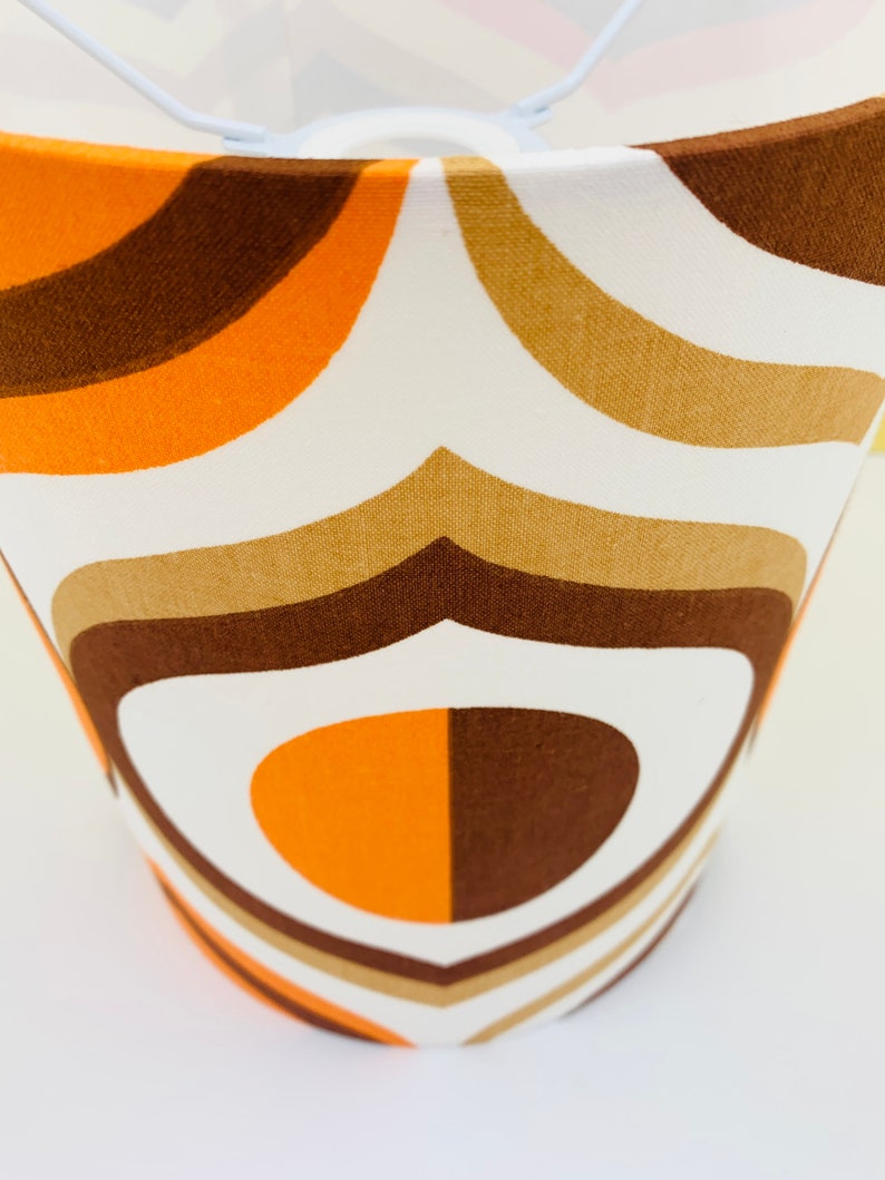 Puede incluir: Pantalla de l&aacute;mpara con dise&ntilde;o retro. La tela presenta un patr&oacute;n repetido de formas curvas en tonos marr&oacute;n, naranja y beige sobre un fondo blanco. La pantalla tiene una estructura met&aacute;lica blanca.