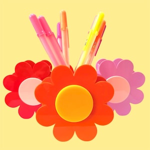 May include: A colourful flower-shaped desk tidy in red, orange, and pink, holding pens and markers. The organiser has a yellow background and a bright design. The pens and markers are in shades of pink, orange, and yellow.