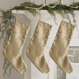 Könnte beinhalten: Drei goldene Weihnachtsstrümpfe mit einem wirbelnden Muster hängen an einem Kamin aus Holz, der mit einer Girlande aus Zedernholz und Holzperlen geschmückt ist.