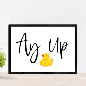 May include: A black framed print with white background and the text "Ay, Up" in black cursive font. A yellow rubber duck is centered below the text.
