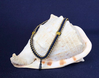 May include: A black and gold braided lanyard with a gold clasp. The lanyard is displayed on a white seashell.