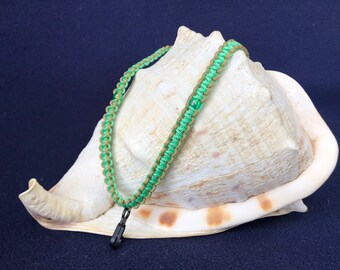 May include: A green and tan braided lanyard with a black clasp. The lanyard is displayed on a large, white seashell against a dark blue background. The lanyard is made of a woven cord.