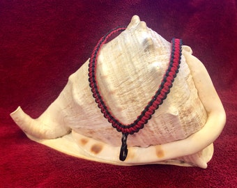 May include: A red and black braided cord necklace with a silver pendant, displayed on a white seashell.