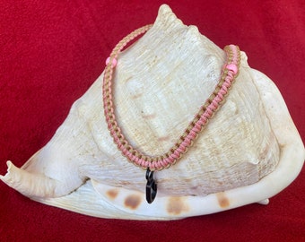 May include: A pink and gold braided cord necklace with a black heart charm. The necklace is displayed on a white seashell.
