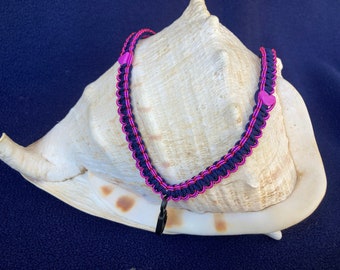 May include: A black and pink braided bracelet with pink heart-shaped beads. The bracelet is on a white seashell.