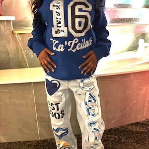 May include: A blue sweatshirt with the number 16 and the text "Sweet 16 Ka'Leilah" in silver glitter. The person is wearing blue jeans with painted designs including a heart, the word "Capricorn", and the number 16.