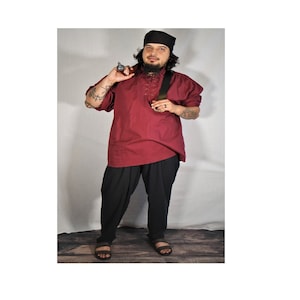 May include: A man wearing a burgundy shirt with a black collar and a black hat. He is wearing black pants and black sandals. He is holding a small object in his right hand.