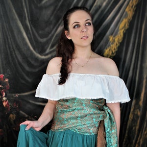 May include: A woman wearing a white off-the-shoulder top with a teal, gold, and brown patterned sash tied around her waist. She is wearing a teal tiered skirt.