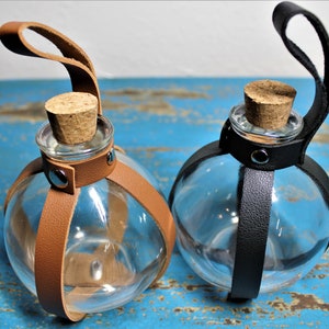 May include: Two clear glass globe-shaped bottles with cork stoppers. One has a tan leather strap and the other a black leather strap. The straps are secured with silver rivets. The bottles are designed to be hung.