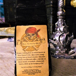 May include: A black bag of coffee with a label featuring a pirate skull and crossbones. The label reads "Rum Runners Isle" and describes the coffee as a non-alcoholic rum flavored coffee. The bag is 8 ounces and is a 1/2 lb drip grind. The label also includes the company name and contact information.