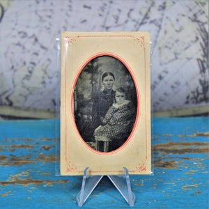 May include: An antique tintype photograph of a woman and a young girl. The woman is wearing a dark dress and the girl is wearing a patterned dress. The photograph is in an oval frame with a red border.