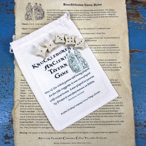 May include: A white cloth bag with a drawstring closure, containing five bone dice. The bag has a brown label with the text "Knucklebones Ancient Tavern Game". The label is surrounded by a green and brown illustration of a tavern scene.