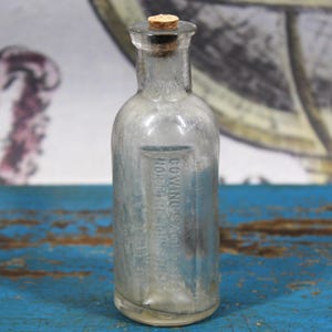 May include: A clear glass bottle with a cork stopper. The bottle has a rectangular label with text. The bottle is on a blue surface. The bottle appears to be an antique.