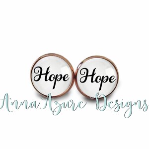 May include: A pair of white glass earrings with a rose gold rim. The earrings feature the word "Hope" in black cursive lettering.