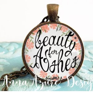 May include: A round glass pendant necklace with a floral design and the text "beauty for ashes" with the scripture reference "Isaiah 61:3".