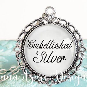 May include: A silver pendant necklace with a round glass charm that says "Embellished Silver". The charm is surrounded by a decorative silver frame.