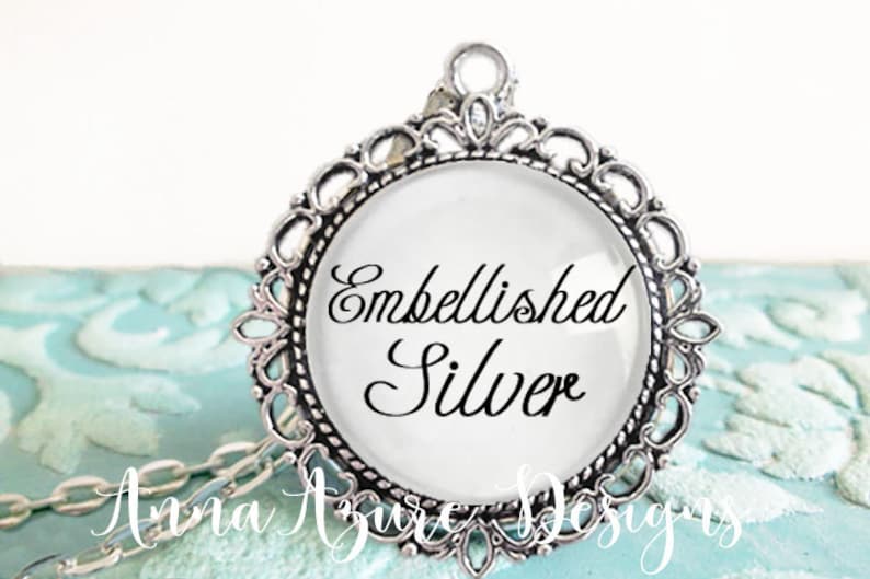 May include: A silver pendant necklace with a round glass charm that says "Embellished Silver". The charm is surrounded by a decorative silver frame.