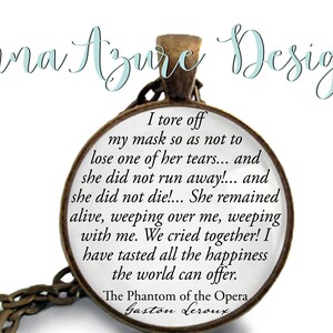 May include: A round pendant necklace with a quote from The Phantom of the Opera by Gaston Leroux. The pendant has a bronze-colored frame and chain. The text is in black on a white background. The brand name "Anna Azure Designs" is at the top.