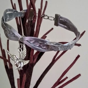 May include: A silver bracelet with a braided leather strap and a silver eagle charm. The bracelet is on a brown twig background.