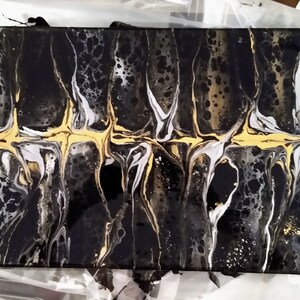 May include: Abstract art painting with black, white, and gold paint. The paint is swirled and layered to create a unique design. The painting is on a canvas.