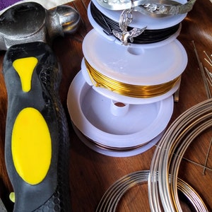 May include: A close-up of a variety of spools of wire, including silver, black, gold, and copper. The spools are stacked on top of each other and are surrounded by a hammer and a set of metal rings.