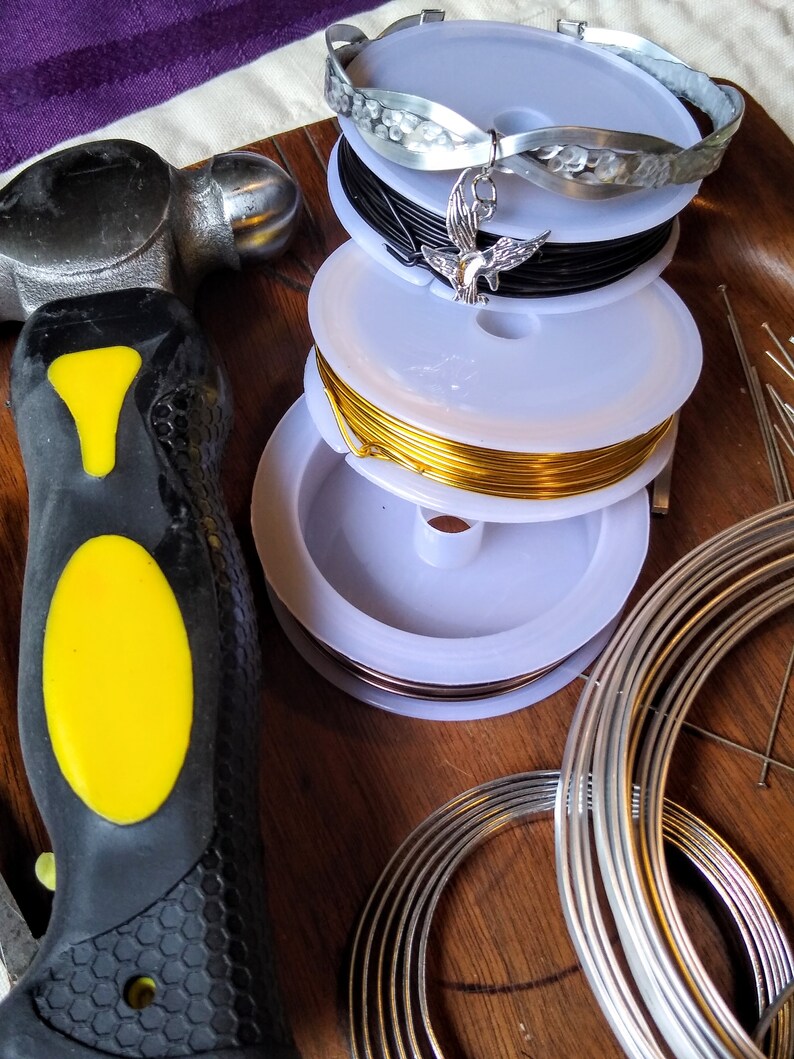 May include: A close-up of a variety of spools of wire, including silver, black, gold, and copper. The spools are stacked on top of each other and are surrounded by a hammer and a set of metal rings.