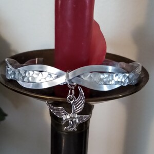 May include: A silver bracelet with a hammered texture and a silver eagle charm. The bracelet is on a candle holder.
