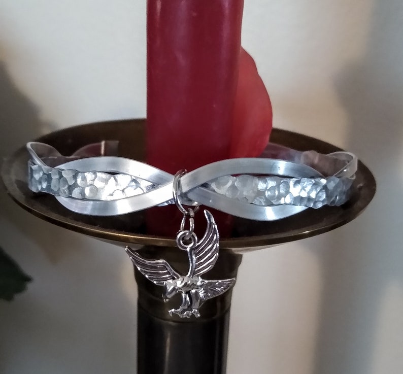 May include: A silver bracelet with a hammered texture and a silver eagle charm. The bracelet is on a candle holder.