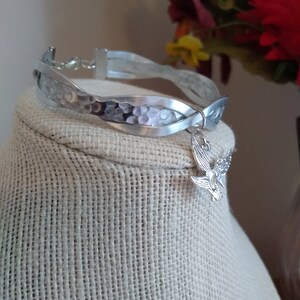 May include: A silver bracelet with a hammered finish and a braided design. The bracelet has a small silver charm in the shape of a dove attached to it.