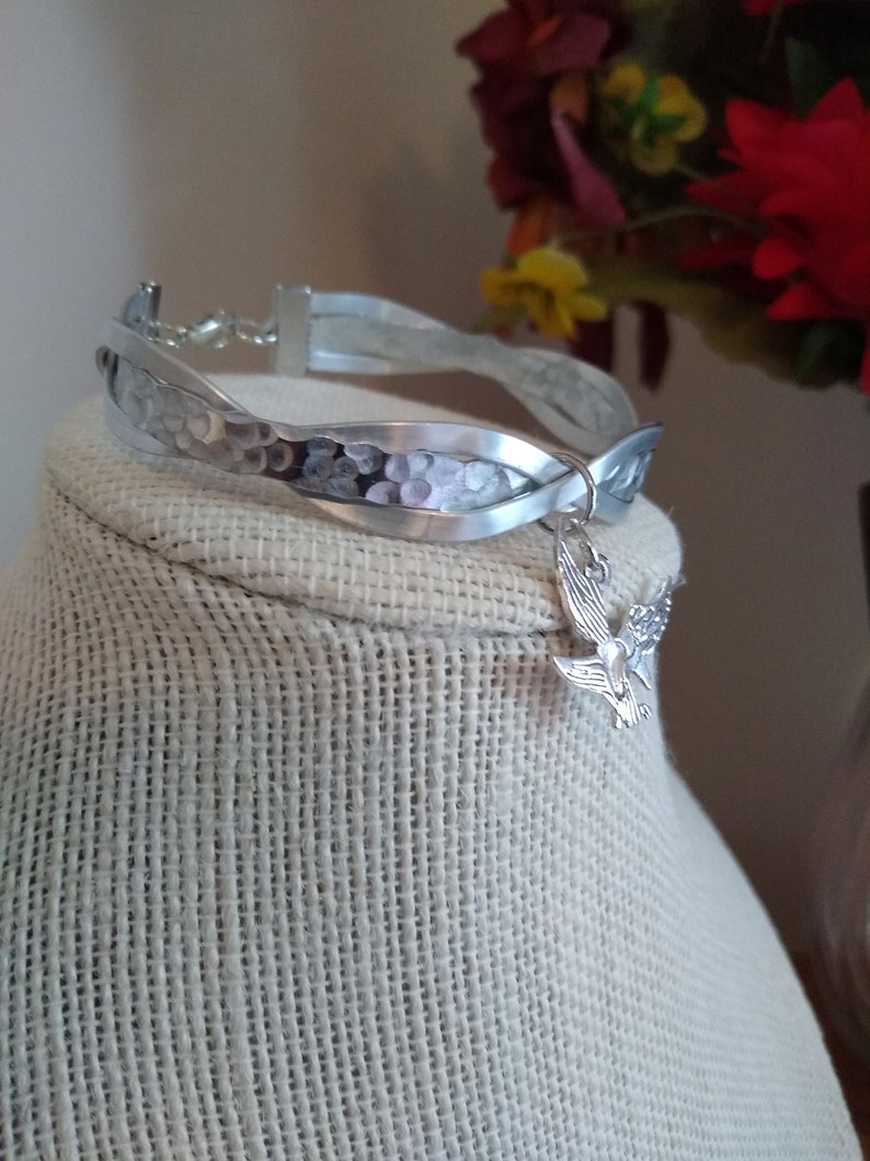May include: A silver bracelet with a hammered finish and a braided design. The bracelet has a small silver charm in the shape of a dove attached to it.