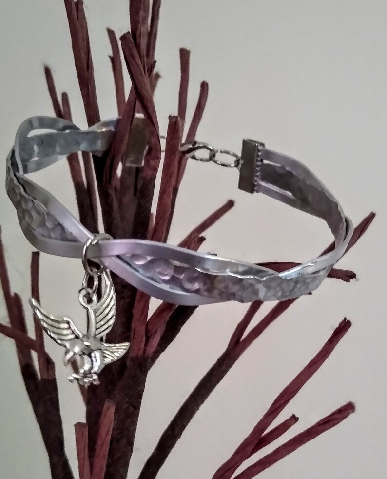 May include: A silver bracelet with a braided leather strap and a silver eagle charm. The bracelet is on a brown twig background.