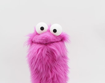 May include: A vibrant, plush puppet in a bright magenta hue. The puppet has large, round white eyes with black pupils and a smiling mouth. The body is cylindrical and covered in long, shaggy fur, creating a playful and whimsical appearance.