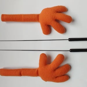 Può includere: Due mani di pupazzo in feltro arancione con aste di metallo nero attaccate sul retro. Le aste sono lunghe circa 30 cm.