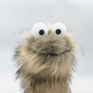 May include: A plush puppet with large, white, googly eyes and black pupils. The puppet's fur is a mix of brown, tan, and gray, creating a textured appearance. The puppet is designed to resemble a friendly monster or creature.